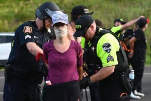 a person in a maroon shirt stares defiantly at the camera as police handcuff her