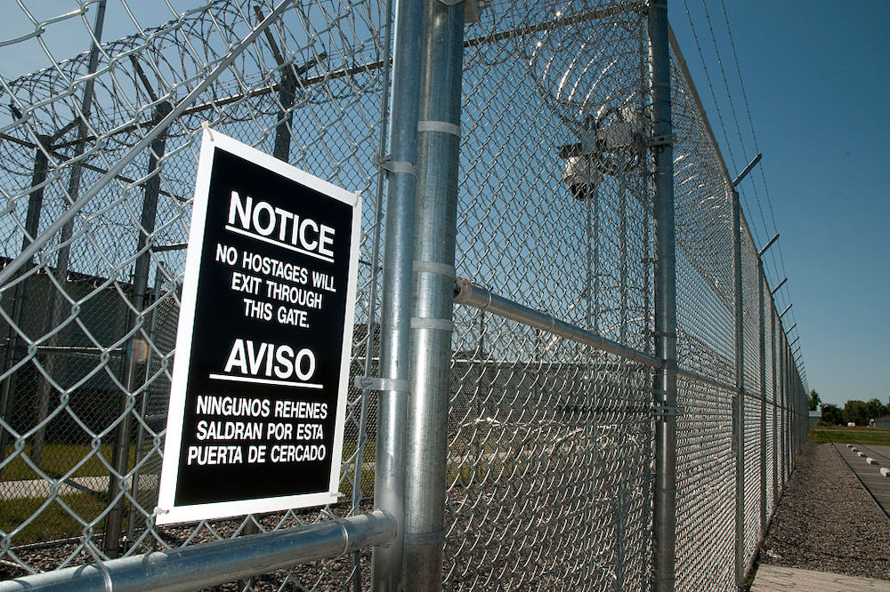 A grim jail fence with razor wire and a sign warning that 'no hostages shall exit through this gate.'